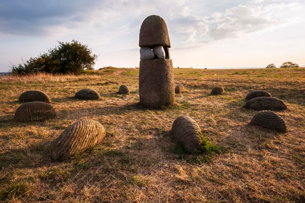 DasBild zeigt eine Landschaft mit trockenen Gräsern, in der mehrere bienenkorbartige Steinstrukturen kreisförmig angeordnet sind. In der Mitte steht eine größere Steinskulptur mit einer abgerundeten Spitze und eingeklemmten Steinen in der Mitte.