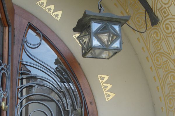 Detail des Eingangsportal am Großen Haus Glückert. Eine kleine Lampe aus Metall und Glas hängt an der Wand. Die Wand ist mit goldenen Ornamenten verziert. Eine Ecke der Runden Holztür ist sichtbar.