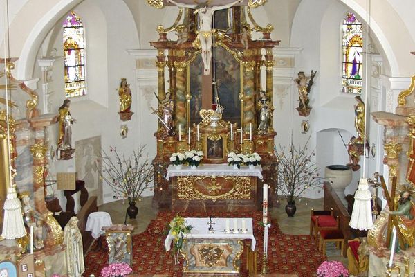 Blick von der Empore auf den Hauptaltar umgeben von einigen Heiligenfiguren, zwei bunten Kirchenfenstern und einem Taufbecken rechts im Altarraum.