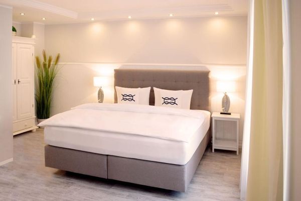 Mehrere Deckenstrahler beleuchten ein Schlafzimmer mit einem grauen Boxspringbett. Daneben steht eine hohe, grüne Pflanze neben einem weißen Kleiderschrank.