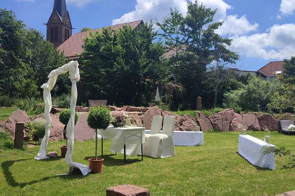Garten, der für eine Hochzeit dekoriert ist. Ein Bogen sowie Bänke und Stühle mit weißen Hussen. Im Hintergrund ist eine Kirche und Bäume zu sehen.