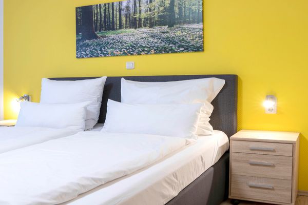 Doppelbett mit weißer Bettwäsche und grauem Kopfteil zwischen zwei Nachttischen mit Leselampen vor einer gelben Wand mit Waldbild.