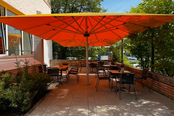 Auf dem Bild ist der Außenbereich zu sehen. Hier befindet sich eine Terrasse, auf welcher mehrere Tische mit Stühlen stehen. Darüber ist ein großer Sonnenschirm aufgespannt.