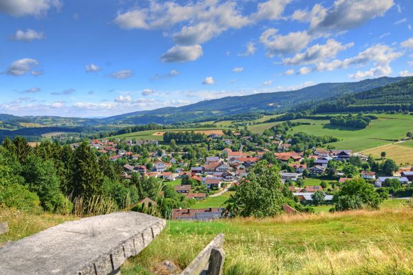 Unter weiß blauem Himmel steht auf einer Wiese ein Holztisch zwischen zwei Bänken. Von hier aus bietet sich ein wunderbarer Blick hinunter auf den Ort Haibach und die umliegenden Wälder.