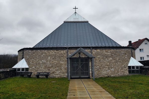 Bei bewölktem Wetter steht die aus Steinen gemauerte Kirche mit Blechdach auf der grünen Wiese am Kirchgelände. Ein Kreuz ziert die Spitze des Daches.