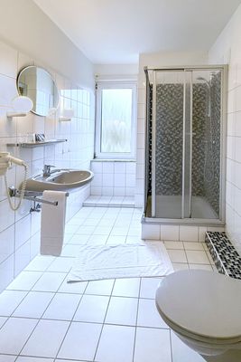 Das Bild zeigt ein modernes Badezimmer mit weißen Fliesen, einem Waschbecken mit Spiegel und Wandlampen, sowie einer Duschkabine mit Glastür. Ein Fenster mit Milchglas sorgt für Tageslicht, und ein Haartrockner sowie Handtücher sind mit Halterungen an der Wand angebracht.