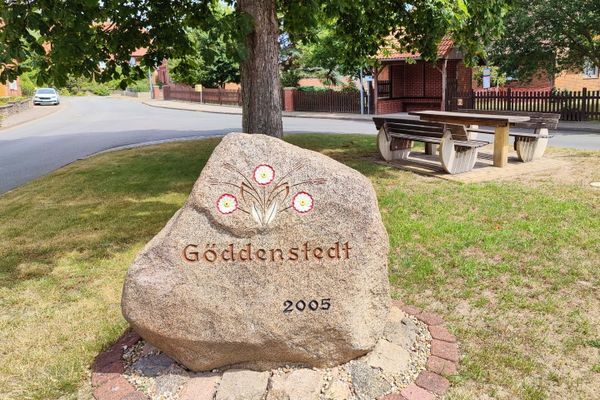 Großer beschrifteter Findling mit der Aufschrift ‚Göddenstedt 2005‘ unter einem Baum auf einer Grünfläche in der Dorfmitte von Göddenstedt, mit Sitzgruppe und Fachwerkgebäude im Hintergrund in der Lüneburger Heide.