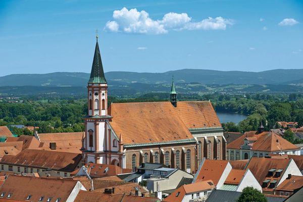 Luftaufnahme der Karmelitenkirche Straubing, umgeben von Dächern angrenzender Gebäude. Im Hintergrund fließt die Donau vor hügeligem, bewaldetem Umland.