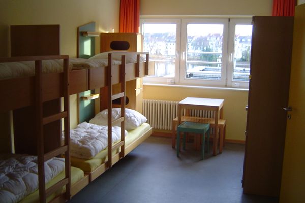 Jugendherbergszimmer mit zwei Etagenbetten, kleinem Tisch mit zwei Hockern und Fenster mit Blick nach draußen