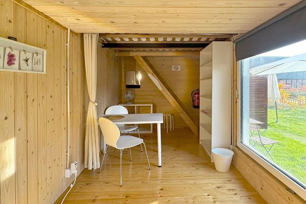 Schmaler Raum mit Holzverkleidung, Tisch mit zwei Stühlen und großem Fenster auf der rechten Seite. Am Ende des Raums stehen ein Regal und weitere Möbel.