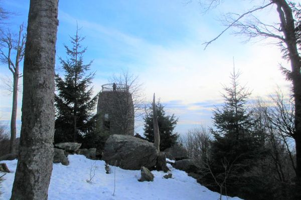 Im Vordergrund liegt eine leichte Schneedecke zwischen einzelnen Bäumen und Felsen; im Hintergrund steht der Hirschenstein-Turm, auf dem eine Person die Aussicht genießt.