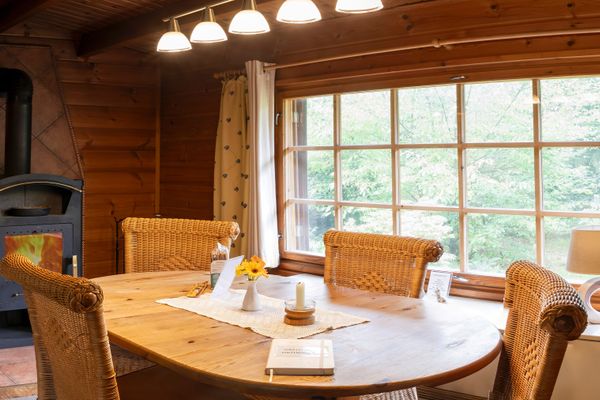 Großer Holztisch mit Stühlen in einem Raum mit Kaminofen und Fenster, beleuchtet durch Deckenlampen.