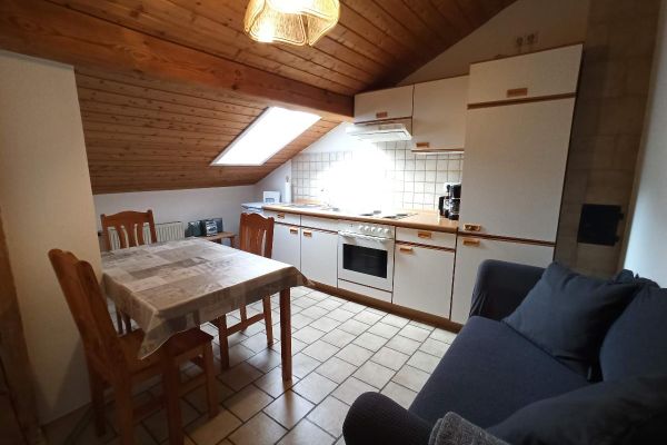 Unter einer holzvertäfelten Dachschräge mit Fenster steht eine helle Küchenzeile. Daneben befinden sich ein Holz-Esstisch mit drei Stühlen und gegenüber ein graues Sofa.