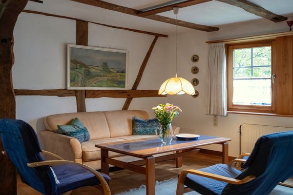 Wohnzimmer mit Sofa, zwei Sesseln, Tisch und Fenster, Holzbalken an der Wand.