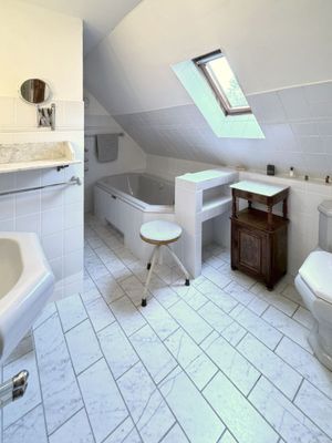 Badezimmer mit Badewanne unter einer Dachschräge, daneben ein Fenster, Fliesenboden und ein kleiner Hocker.