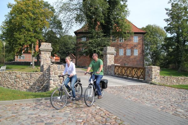 Zwei Radfahrende fahren vor einer Steinbrücke und den Gebäuden der Burg Bodenteich.