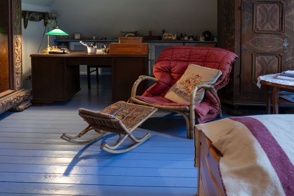 Raum mit gepolstertem Sessel, kleinem Hocker, Schreibtisch mit Lampe und großem Holzschrank.
