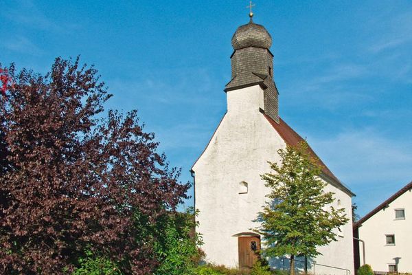 Kleine Kirche mit schindelbedecktem, mehreckigem Kirchturm und Zwiebelkuppel. Links ein blühender Strauch, rechts ein Laubbaum, dahinter ein Gebäude.