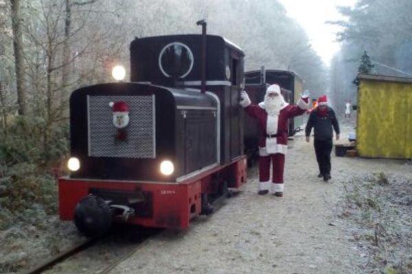 Die alte Lok im winterlichen Wald mit Schnee. Die Bahn ist Weihnachtlich geschmückt und neben ihr läuft ein Weihnachtsmann.