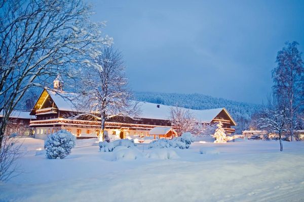 Wunderschön liegt das Hotel traumhaft verschneit und hell erleuchtet in der Abenddämmerung. Auch der Wald im Hintergrund ist tief verschneit.