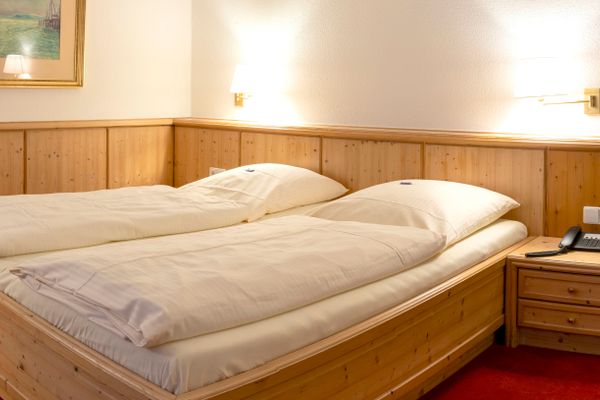 Das Bild zeigt ein gemütliches Schlafzimmer mit einem Doppelbett aus Holz, weißen Bettdecken und Kissen, Holzpaneelen an der Wand und einem gerahmten Bild darüber. Auf beiden Seiten des Bettes stehen Nachttische, einer mit Lampe und Telefon. Ein roter Teppich bedeckt den Boden.