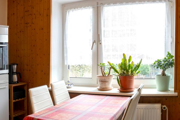 Das Bild zeigt eine Küche mit einem Esstisch, der mit einer rot-gemusterten Tischdecke bedeckt ist. Links im Bild ist ein weißer Küchenschrank mit Backofen und Mikrowelle darauf. Eine Kaffeemaschine steht daneben. Rechts sind zwei große Fenster mit weißen Gardinen und Topfpflanzen zu sehen. Die Wände sind holzverkleidet.