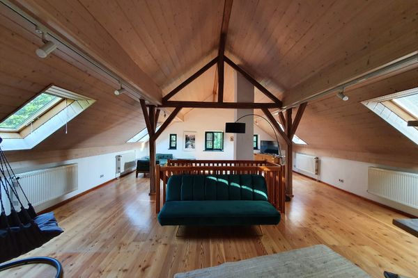 Großer Wohnbereich im Dachgeschoss mit mehreren Fenstern und Dachfenstern. Holzdecke und Holzboden, grüne Sofas und ein Hängesessel mit Gestell.