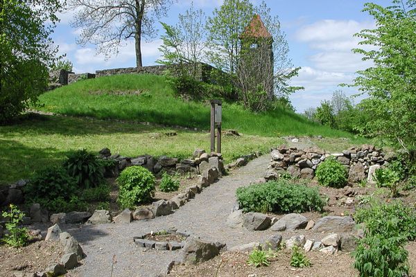 Ein kleiner Schotterweg, gesäumt von Basaltsteinen, führt durch ein Kräuterbeet. Auf den Parzellen sind allerlei Pflanzen zu sehen. Eine Schautafel steht dahinter. Weiter hinten sind Bäume auf einer hanglagigen Wiese zu sehen und auf dem Bergsporn ein Bergfried mit rot eingedecktem Dach sowie Mauer ringsum.