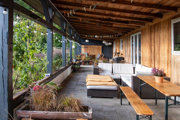 Eine überdachte, gepflasterte Terrasse mit einer holzverkleideten Hauswand rechts und dunklen Holzpfeilern links, die den Blick in einen grünen Garten freigeben. Auf der Terrasse stehen ein dunkles Ecksofa mit weißen Polstern, ein Holztisch mit Holzbank sowie bepflanzte Blumenkübel.