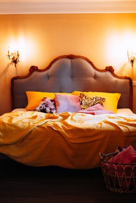 Auf dem Bild ist ein Bett in einem Zimmer zu sehen. Am Bett befindet sich eine barocke Bettlehne. Daneben hängen an der Wand Beleuchtungen in Form von Kerzenhaltern.
