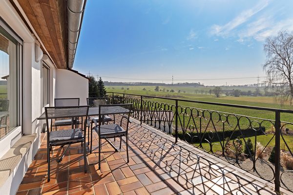 Terrasse der Ferienwohnung Bergfeld mit einem Tisch und 4 Stühlen; Ausblick auf eine weitflächige Landschaft mit blauem Himmel