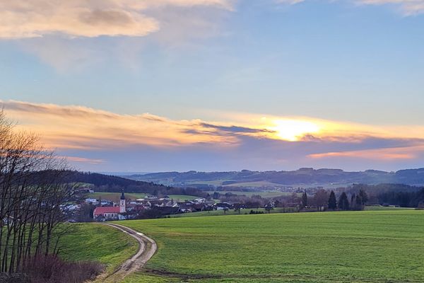 Im Vordergrund eine große Wiese, durch die ein Weg zum Ort Neukirchen führt, in dessen Mittelpunkt die Kirche steht. Im Hintergrund etwas verstreut einige Weiler und eine weite Hügellandschaft.