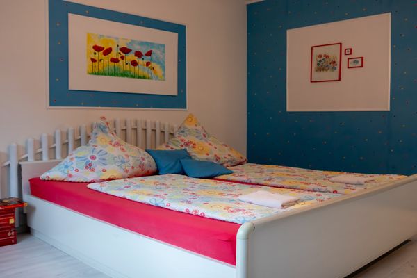 Ein weißes Doppelbett mit einem pinkfarbenen Spannbettlaken und bunter Bettwäsche mit Blumenmuster. Darauf liegen zwei blaue Dekokissen und gefaltete Handtücher. Die Wand hinter dem Kopfende ist weiß mit einem großen Bild von roten Mohnblumen auf blauem Grund. Die rechte Wand ist blau mit bunten Punkten und drei kleinen gerahmten Bildern. Links neben dem Bett steht ein Stapel Bücher auf dem Boden.
