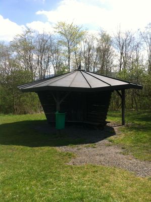 Der Wanderer steht vor einem Unterstand mit ausladendem Dach welches sich auf einer Wiese vor einem Laubbaumwald befindet.
