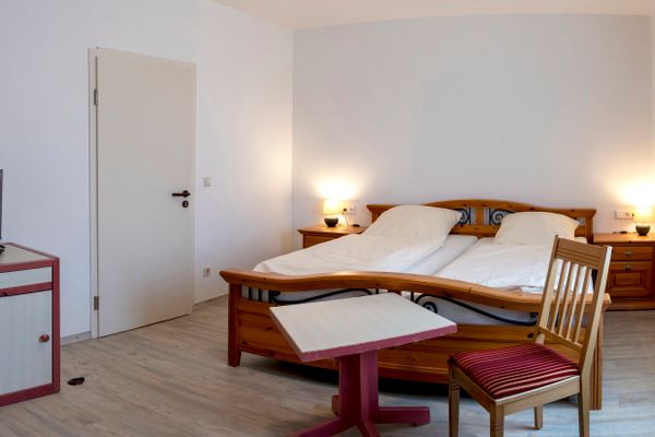 Das Bild zeigt ein gemütliches Schlafzimmer mit einem Holz-Doppelbett, das mit weißen Bettdecken und Kissen ausgestattet ist. Neben dem Bett stehen zwei Nachttische mit Lampen, und im Raum befinden sich ein Schrank mit einem kleinen Fernseher sowie ein Tisch mit einem Stuhl.