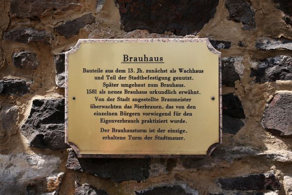Messingschild an einer Basaltsteinwand mit der Überschrift Brauhaus. Weiter steht folgender Text geschrieben: Bauteile aus dem 13. Jahrhundert zunächst als Wachhaus und Teil der Stadtbefestigung genutzt. Später umgebaut zum Brauhaus. 1581 als neues Brauhaus urkundlich erwähnt. Von der Stadt angestellte Braumeister überwachten das Bierbrauen, das von den einzelnen Bürgern vorwiegend für den Eigenbedarf praktiziert wurde. Der Brauhausturm ist der einzige erhaltene Turm der Stadtmauer.