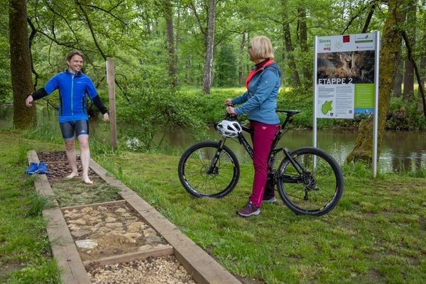 Ein Mann der einen Barfußpfad entlang geht. Neben ihm steht eine Frau mit Fahrrad.