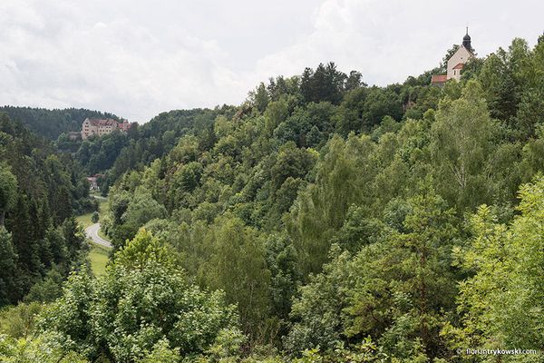 Blick auf ein grünes Tal, oberhalb dessen sich eine Burg im Hintergrund und eine Kapelle im Vordergrund erheben.