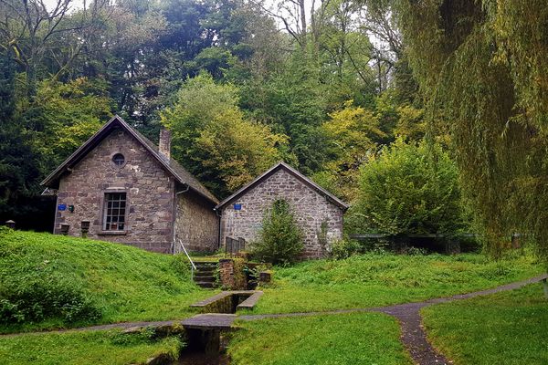 Zwei Häuser aus Basaltsteinen gebaut, sind das zentrale Bildmotiv. Davor geht durch die grüne Wiese ein offener Wasserkanal über den ein Weg führt. Hinter den Häusern ist ein Wald.