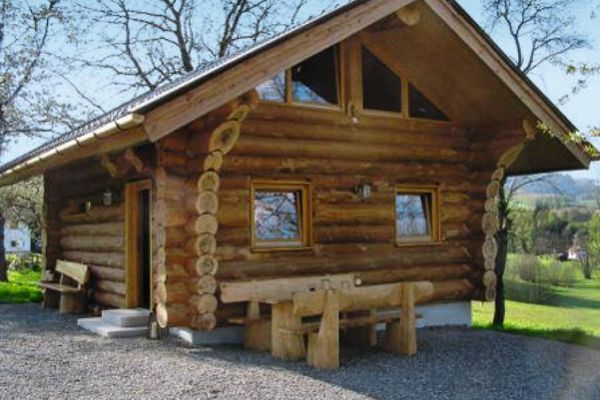 Naturstammhaus mit Giebelfenstern. Auf der kiesbedeckten Fläche vor dem Haus stehen rustikale Holzsitzbänke und ein Tisch.