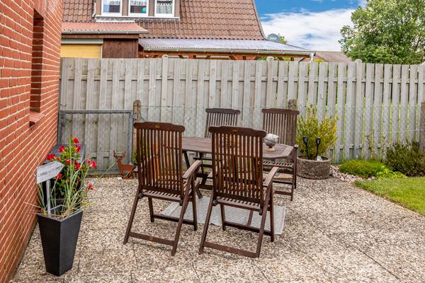 Das Bild zeigt eine Terrasse mit Holzmöbeln bestehend aus 4 Stühlen und einem Tisch auf Steinplatten, umgeben von einem hohen Holzzaun. Pflanzen, ein Blumentopf mit roten Blüten und ein Hausdach sind im Hintergrund.