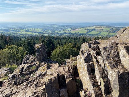 Vom schroffen Basaltfelsen des Geotopes Bilstein im hessischen Vogelsberg geht der Blick weit über das sanft geschwungene Land der Vulkanregion Vogelsberg zum Horizont. Im Gegensatz zum braun-grauen Basaltgestein des Berges Bilstein ist die Landschaft sommerlich in sattes Grün gehüllt und die Felder, Wiesen und Wälder sowie der blaue Himmel wirken sehr einladend.