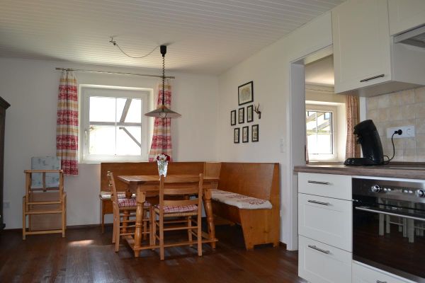 Zwischen Küchenzeile und Essbereich im Landhausstil befindet sich der Durchgang zum Wohnzimmer. Neben dem weißen Sprossenfenster mit halbhohen, rotgemusterten Vorhängen steht ein Kinderhochstuhl. An der Wand hängen mehrere Bilder.