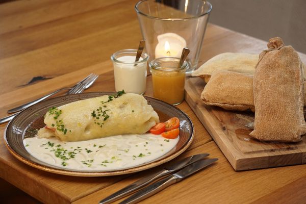 Ein länglicher Kartoffelkloß mit Fleischeinlage liegt mittig zwischen dunkelbrauner Zwiebelsauce rechts und heller cremiger Sauce links auf einem braunen Teller. Der Teller mit Besteck steht auf einem hellbraunen Holztisch. Dahinter liegt ein Holzbrett mit drei länglichen und leicht bräunlichen Leinensäcken übereinandergestapelt. Dazwischen stehen zwei kleine Gläser mit Löffel und cremiger, weißer Füllung sowie Apfelmus vor einem Glas mit einer Kerze.