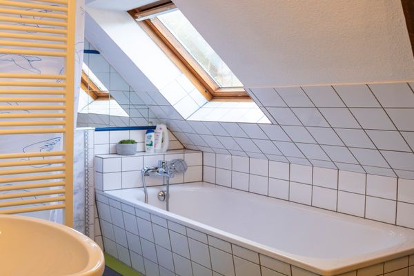 Ein Badezimmer unter einer weißen Dachschräge, in die ein hölzernes Dachfenster eingelassen ist. Im Zentrum befindet sich eine weiß geflieste Badewanne mit silberner Armatur. Im linken Vordergrund sind ein angeschnittenes Waschbecken, ein weißer Sprossenheizkörper und ein Spiegel zu erkennen, während am rechten Rand ein dunkler Holzbalken verläuft.