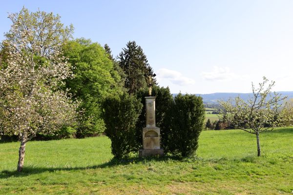 Ein Feldkreuz auf einem Sockel steht, von Bäumen geschützt, auf einer Wiese. Dahinter und daneben weitere Bäume; im Hintergrund Wiesen umgeben von einer bewaldeten Hügellandschaft.
