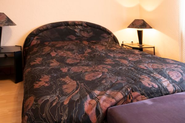 Das Bild zeigt ein Schlafzimmer mit einem Doppelbett, das mit einer dunklen Bettdecke mit floralen Mustern bedeckt ist. Auf beiden Seiten des Bettes befinden sich eingeschaltete Nachttischlampen. Der Boden ist aus hellem Holz.
