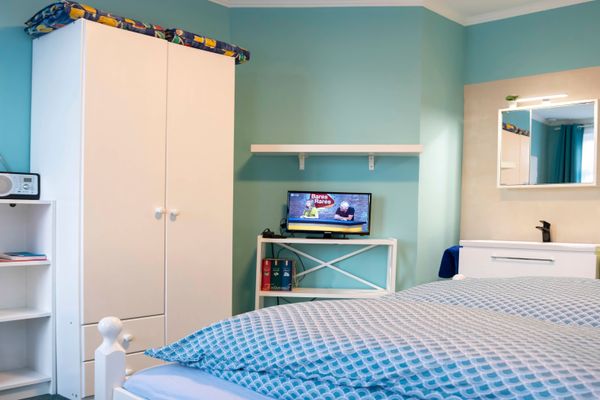 Helles Zimmer mit blauen Wänden. Links steht ein weißer Kleiderschrank mit Regal. Mittig ein Fernseher auf einem weißen Tischchen. Rechts eine Waschecke mit Spiegel. Im Vordergrund ein Bett mit blau gemusterter Bettwäsche.