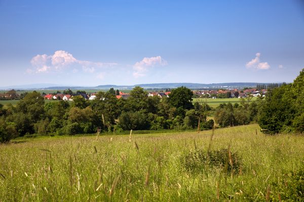 Blick über eine Wiese hinweg auf einen kleinen Ort hinter einer Baumreihe. Dahinter ist weite Landschaft zu sehen. Der Himmel ist strahlend Blau mit wenigen Wolken.