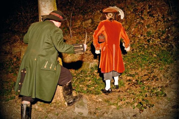 In einem Wald geht ein Mann mit knielanger, roter Jacke und Dreispitz, eine Holzkassette unter dem Arm. Hinter ihm folgt ein Mann in langer, grüner Jacke und mit Hut, der einen Dolch in der Hand hält.